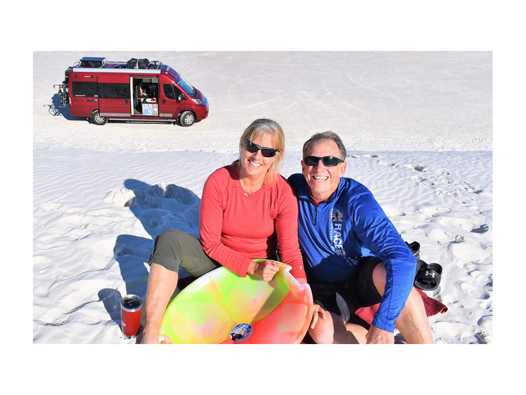 Jim and Rhonda Kohnke sitting in sand at White Sands National Park with red Travato behind them