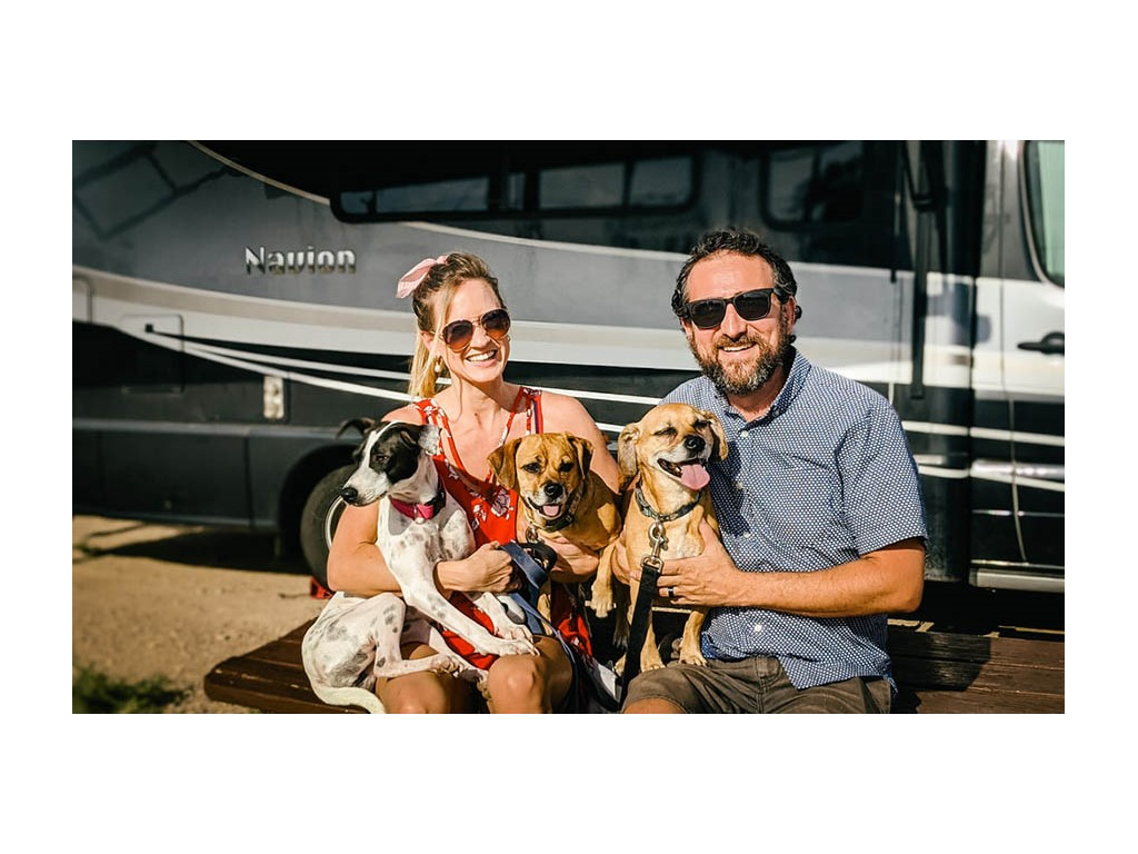 Katelyn and Howard Newstate sitting on picnic table holding three small dogs in front of Winnebago Navion.