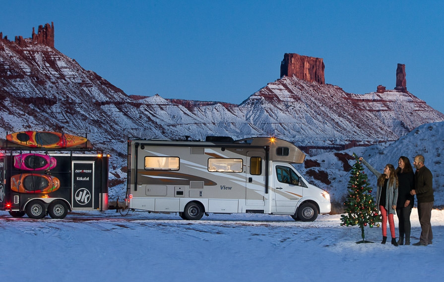 Winnebago View with trailer attached parked on snow covered ground with rock formations in the background and family next to lit Christmas tree out front.