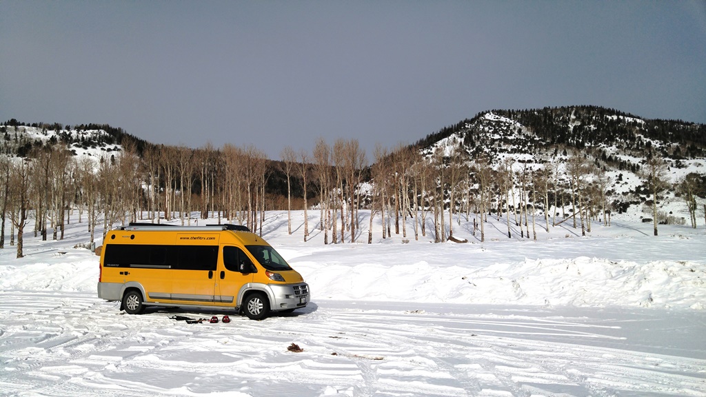 Yellow Winnebago Travato parked on snow covered road with hills behind.