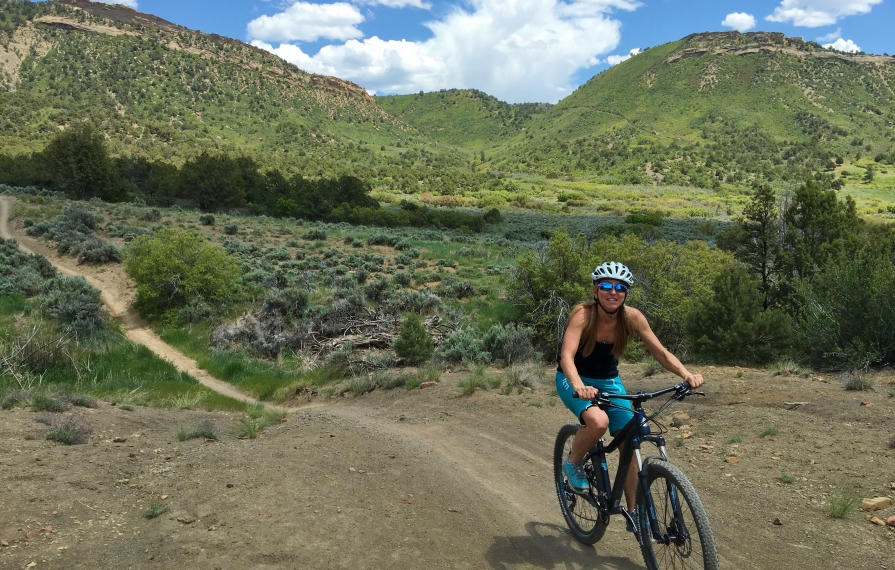 Stef riding bike along a dirt trail with hills in the background.