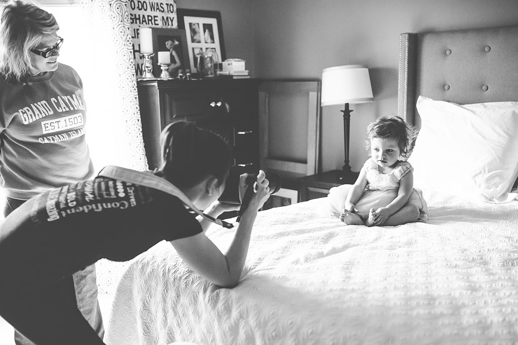 Woman taking a photo of young child sitting on a bed.