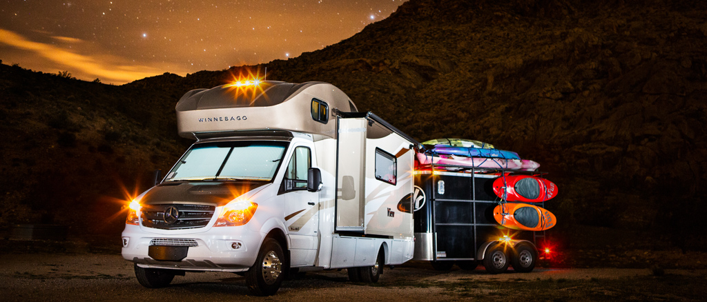 Winnebago View with trailer attached parked near base of a rocky hillside and stars shining in the sky above.