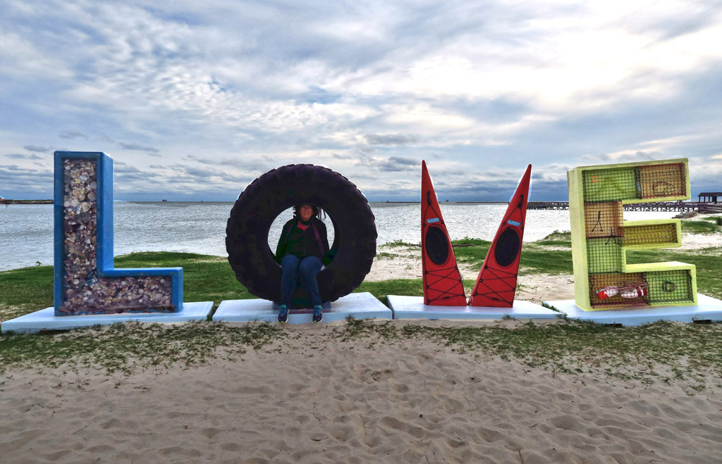 Woman sitting in a large tire used as the "O" in the word "LOVE" spelled out on the beach with different items.