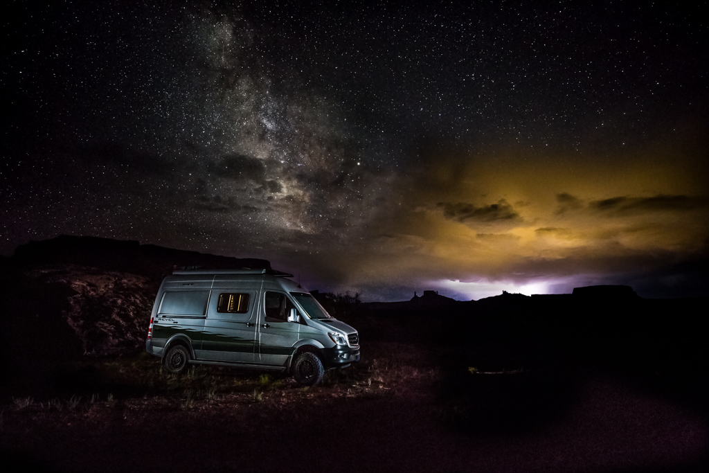 Winnebago Revel parked alone in desert landscape with the Milky Way bright overhead in the nightsky on the left and an approaching storm lighting the sky on the right.