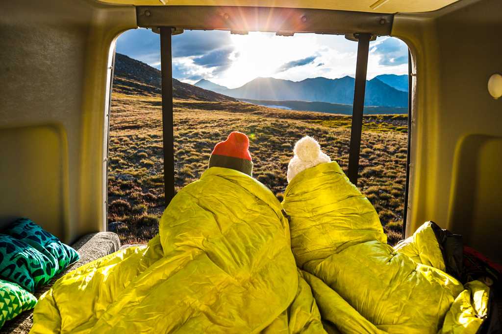 Couple huddled together on the back bed of the Winnebago Revel looking out over the mountains as the sun rises.