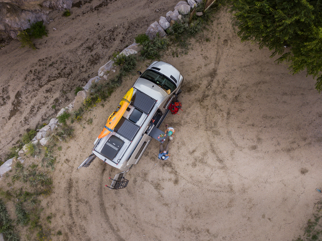 Overhead view of Winnebago Revel with solar panels and a kayak on top parked on dirt ground with Kathy and Peter sitting at the table outside.