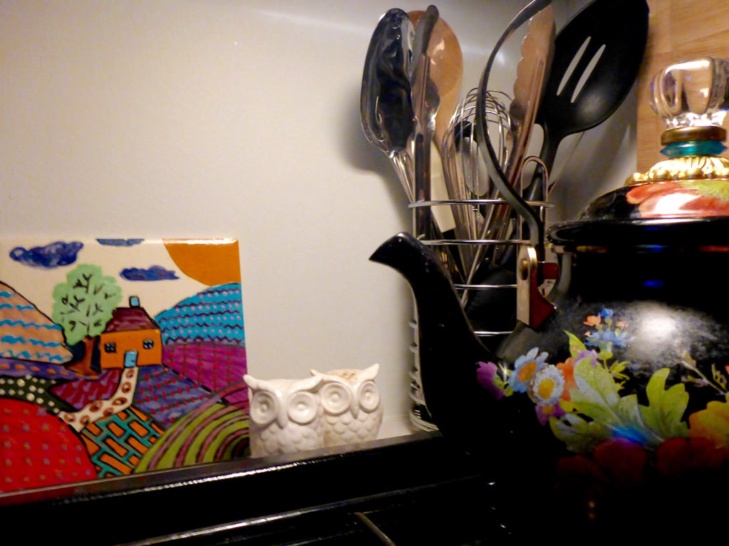 Kitchen utensils, a tea pot and artwork near stovetop.
