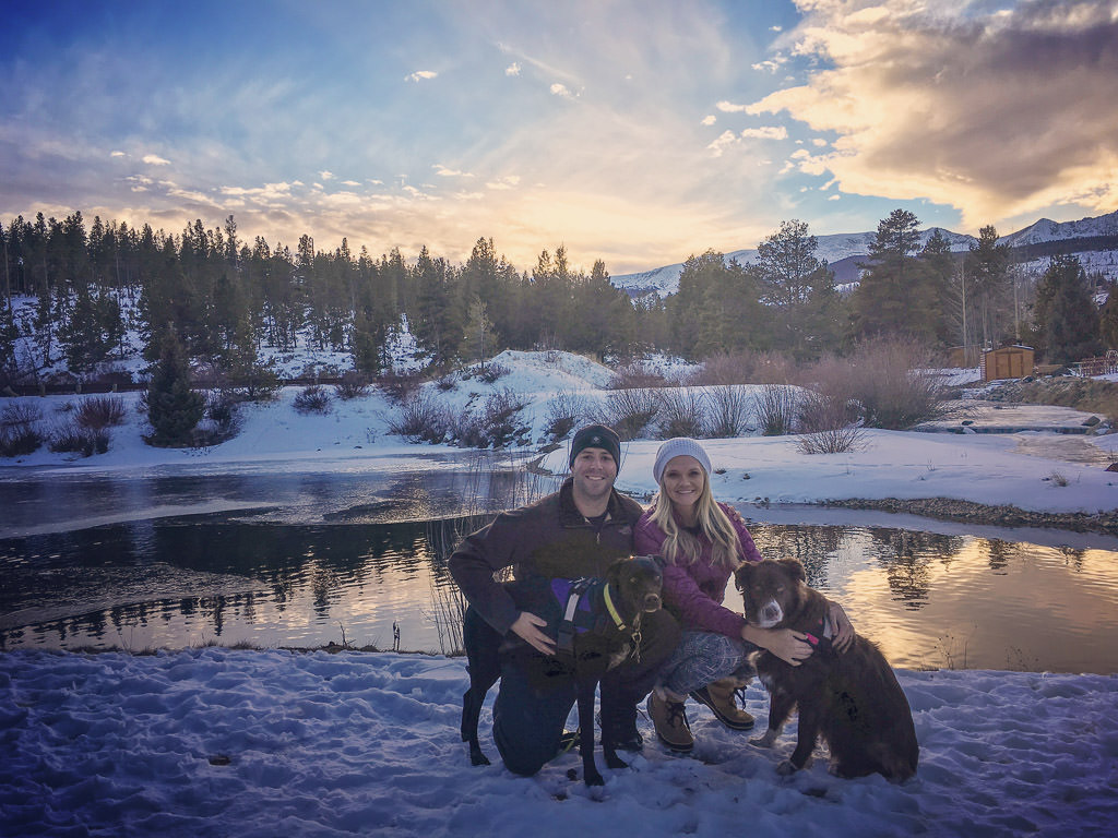 Couple kneeling with their two dogs by a pond with snow covered ground and colorful sky above