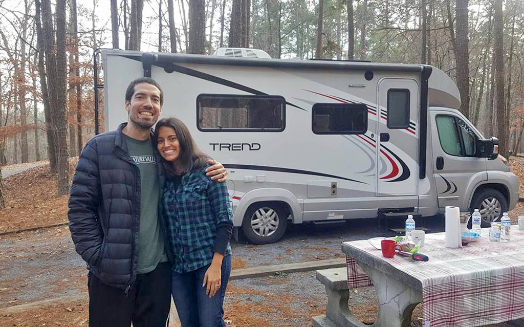 Jon and Nadia standing outside their Winnebago Trend at a tree surrounded campsite.
