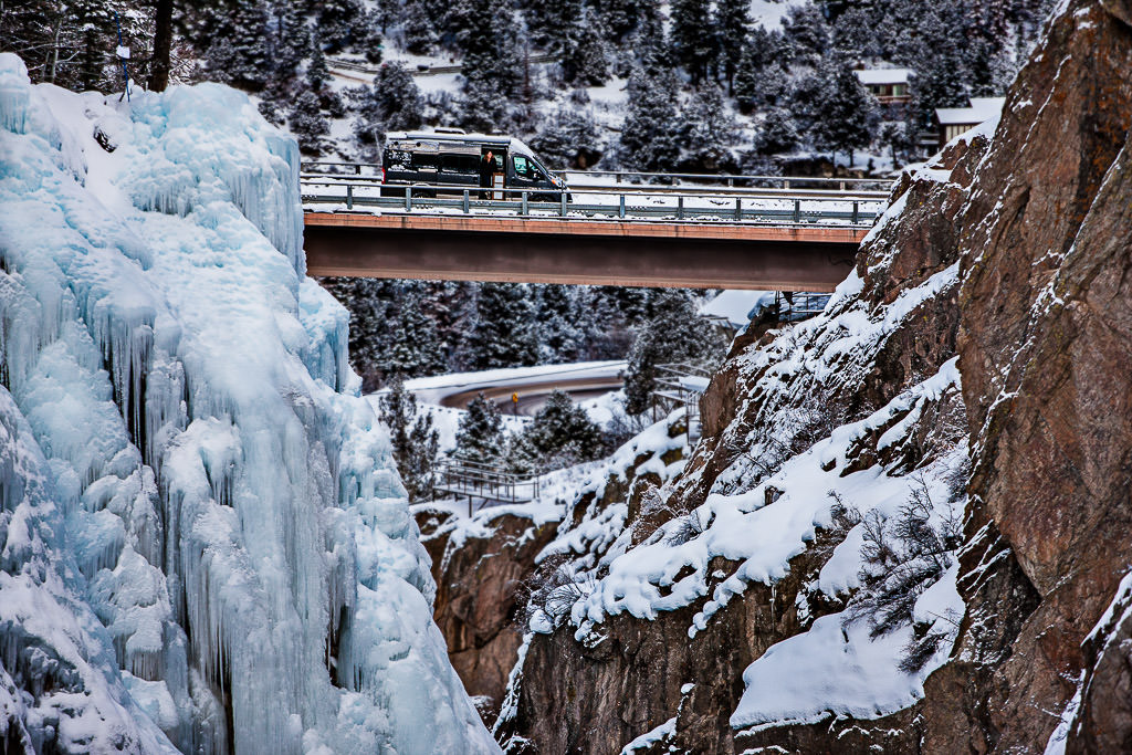 RV parked on a bridge with landscape of ice, snow covered cliffs and trees on either side..