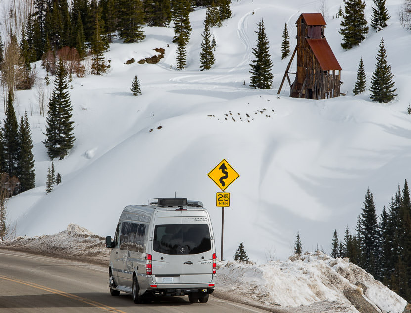 Winnebago Era driving past winding road 25 mph sign with snow covered hillside and evergreens ahead.