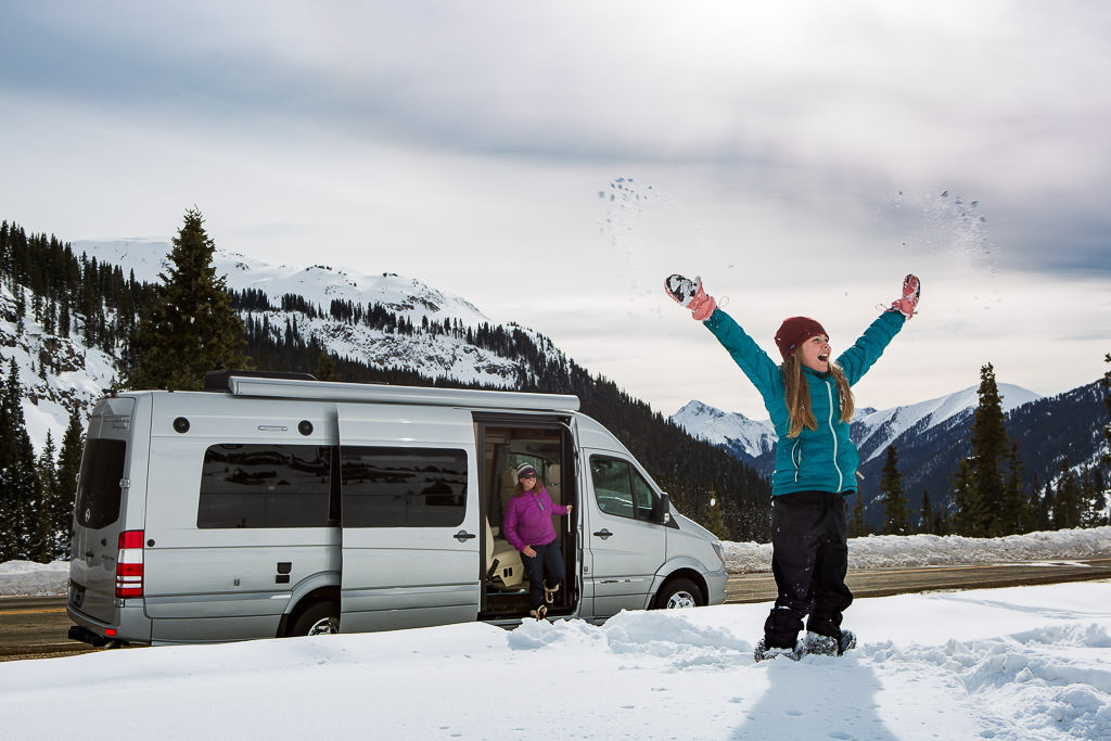 Van parked with snow and tree covered mountains in background, a woman getting out of the van, and a young girl standing in snow throwing snow in the air.