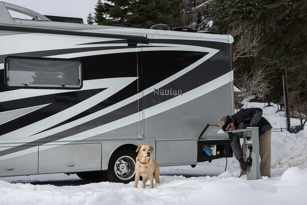Winnebago Navion parked on snow covered ground with dog outside exploring and man plugging unit in to the electric hook-up.