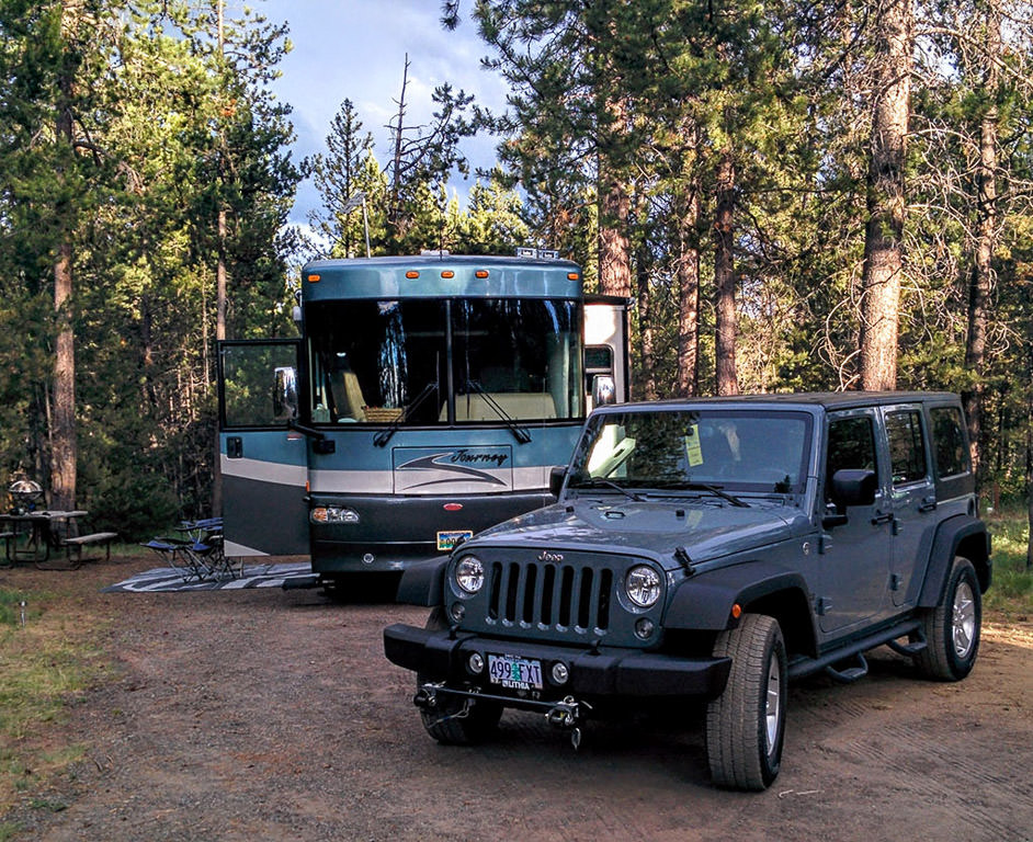 Winnebago Journey and Jeep parked in campsite surrounded by trees.