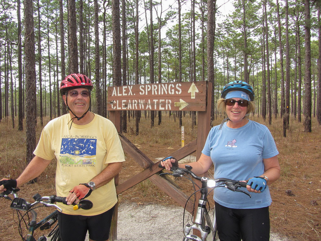 Two people standing with their bikes in front of a sign on the trail for Alex Springs and Clearwater.