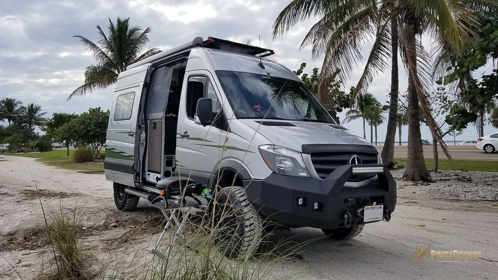 Winnebago Revel parked with side door open on the sandy beach with palm trees in the background.